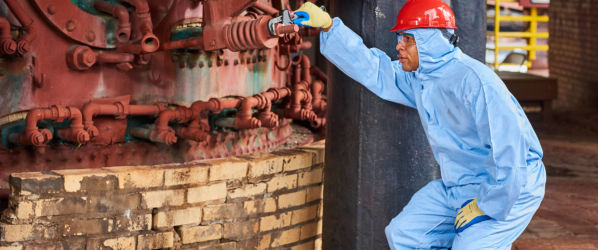 Worker wears Pyrolon Plus 2 Coverall and hard hat while inspecting machinery in a warehouse.